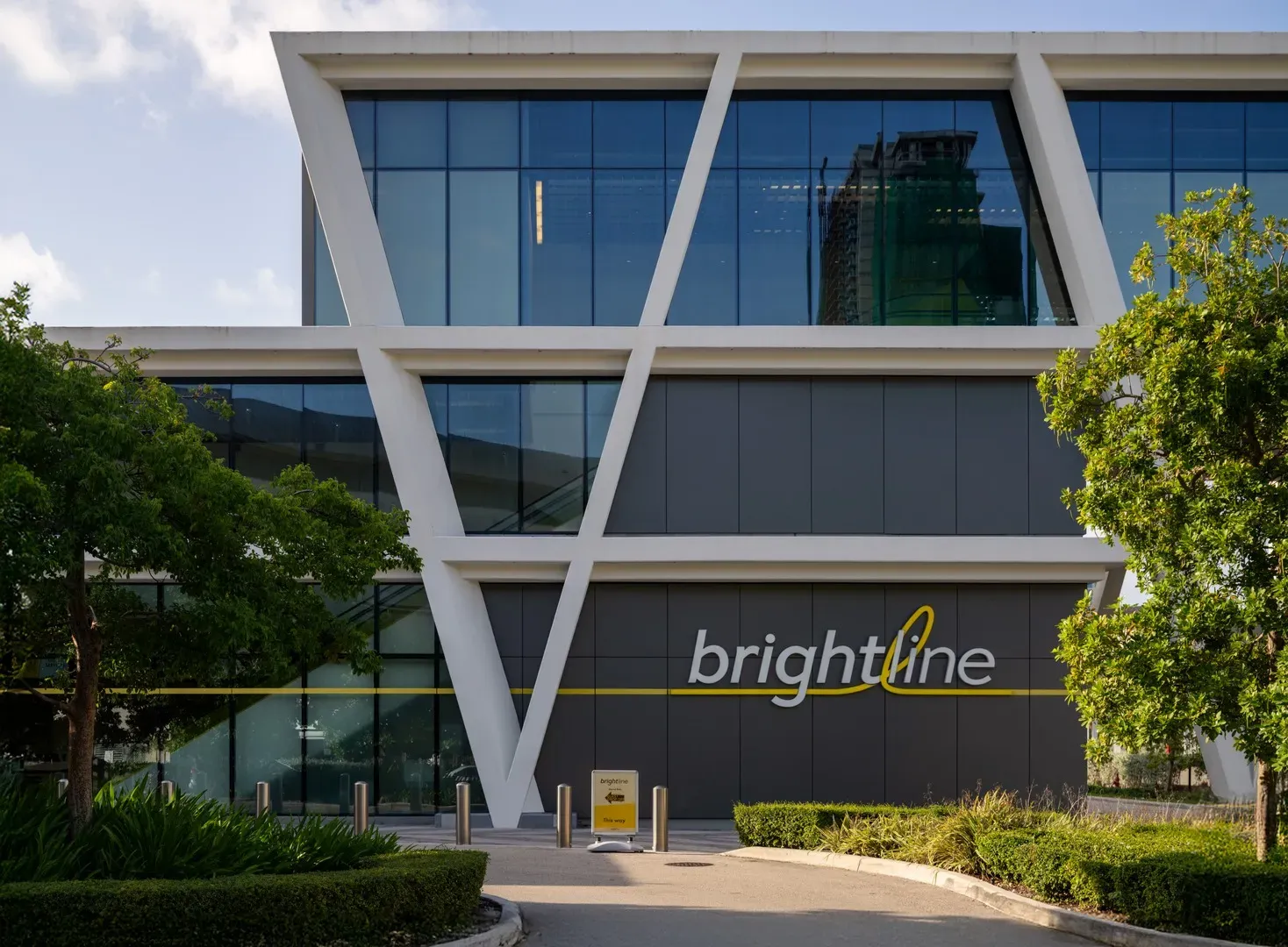 Modern multi-story building with a white geometric facade and a dark gray lower level featuring large reflective windows, framed by green trees.