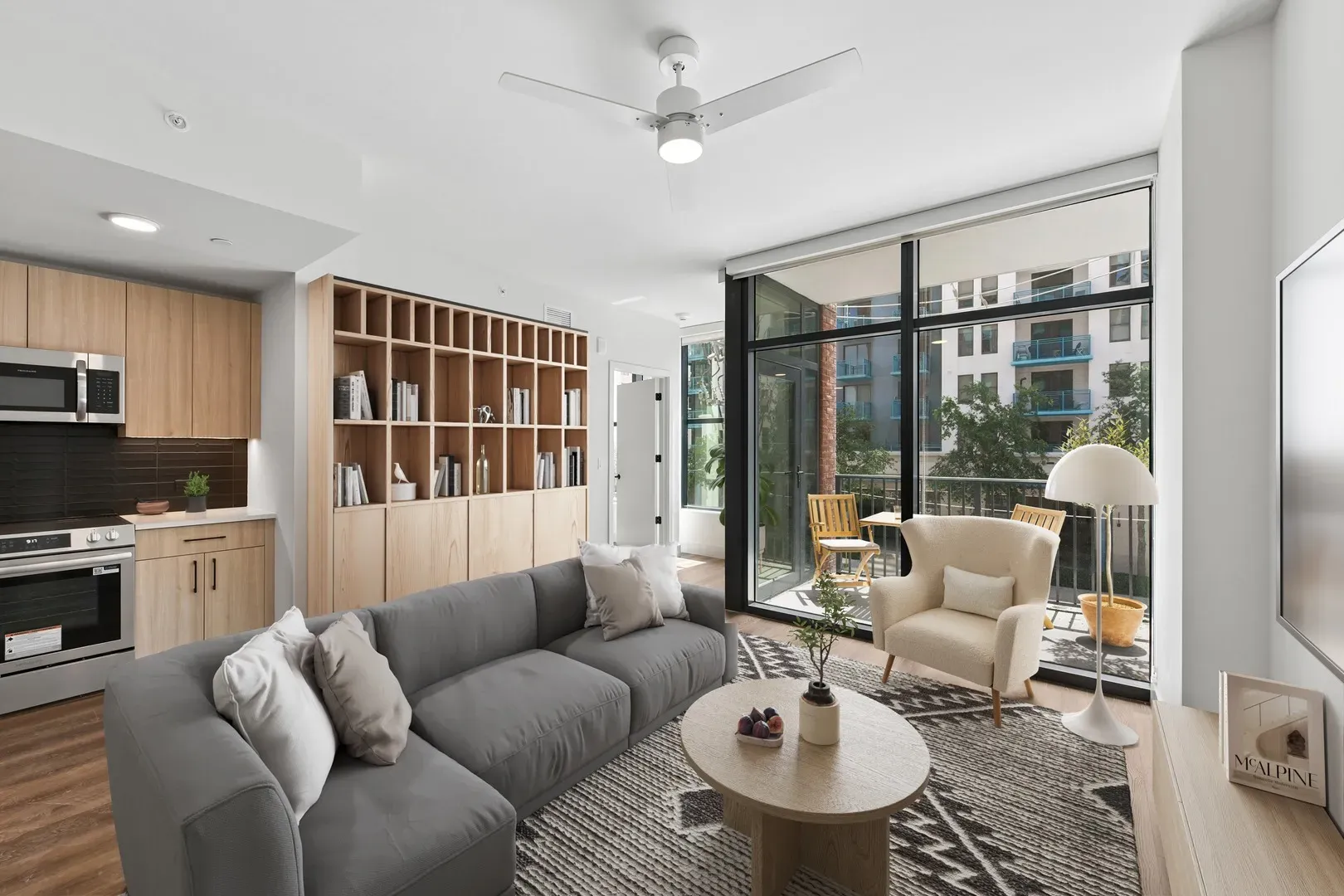 Spacious open-concept living room with a gray sectional sofa, oval coffee table, and kitchen area, opening to a balcony with natural light.