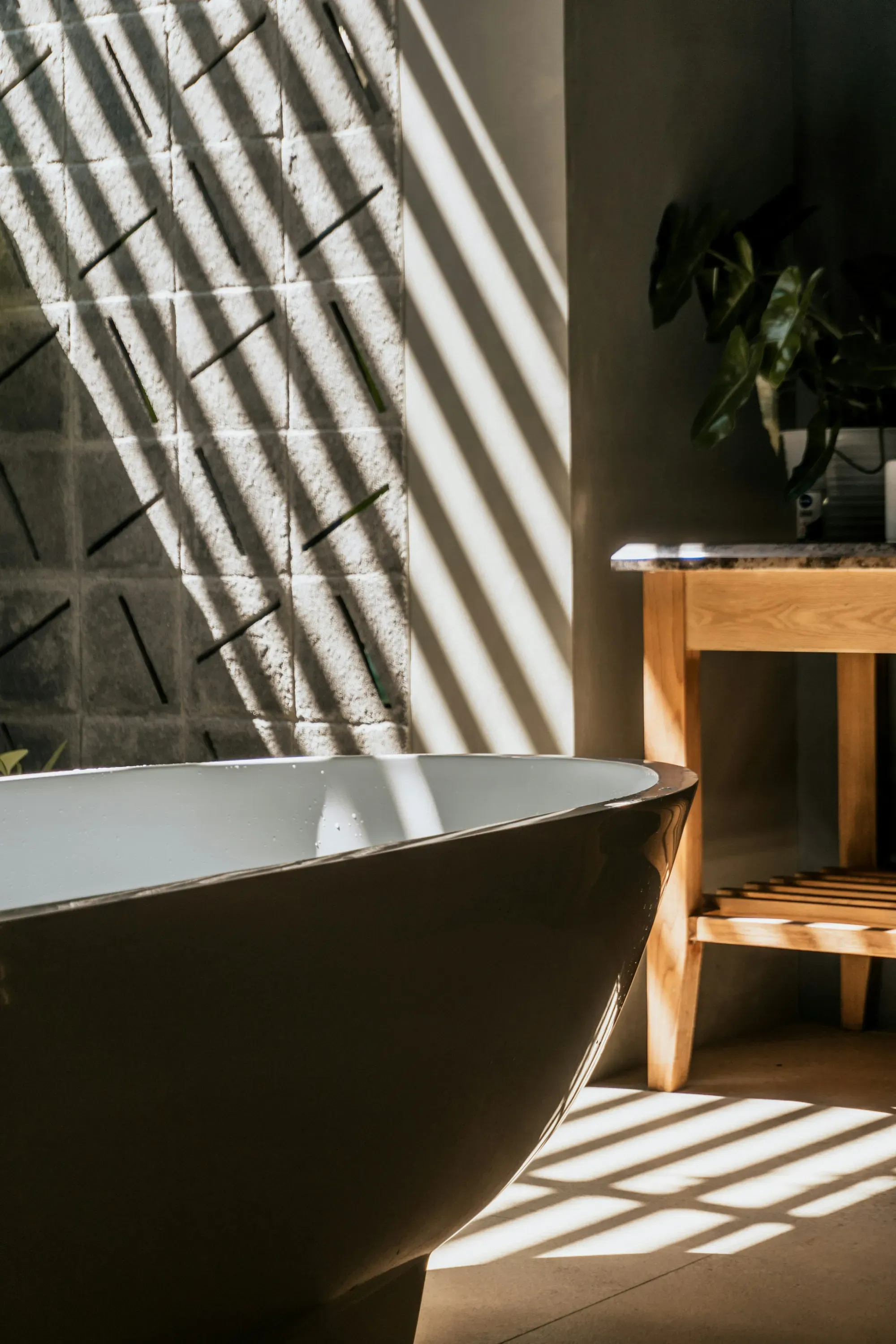Modern bathroom with a dark freestanding bathtub, sunlight casting shadow stripes across the tiled wall, and a potted plant nearby.