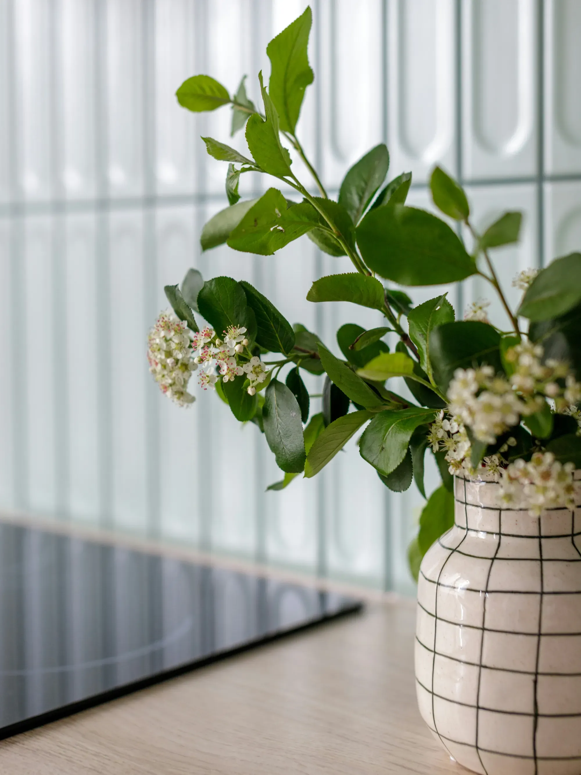 White vase with black grid pattern holding a green plant with white flowers, set on a light countertop against a tiled kitchen wall.