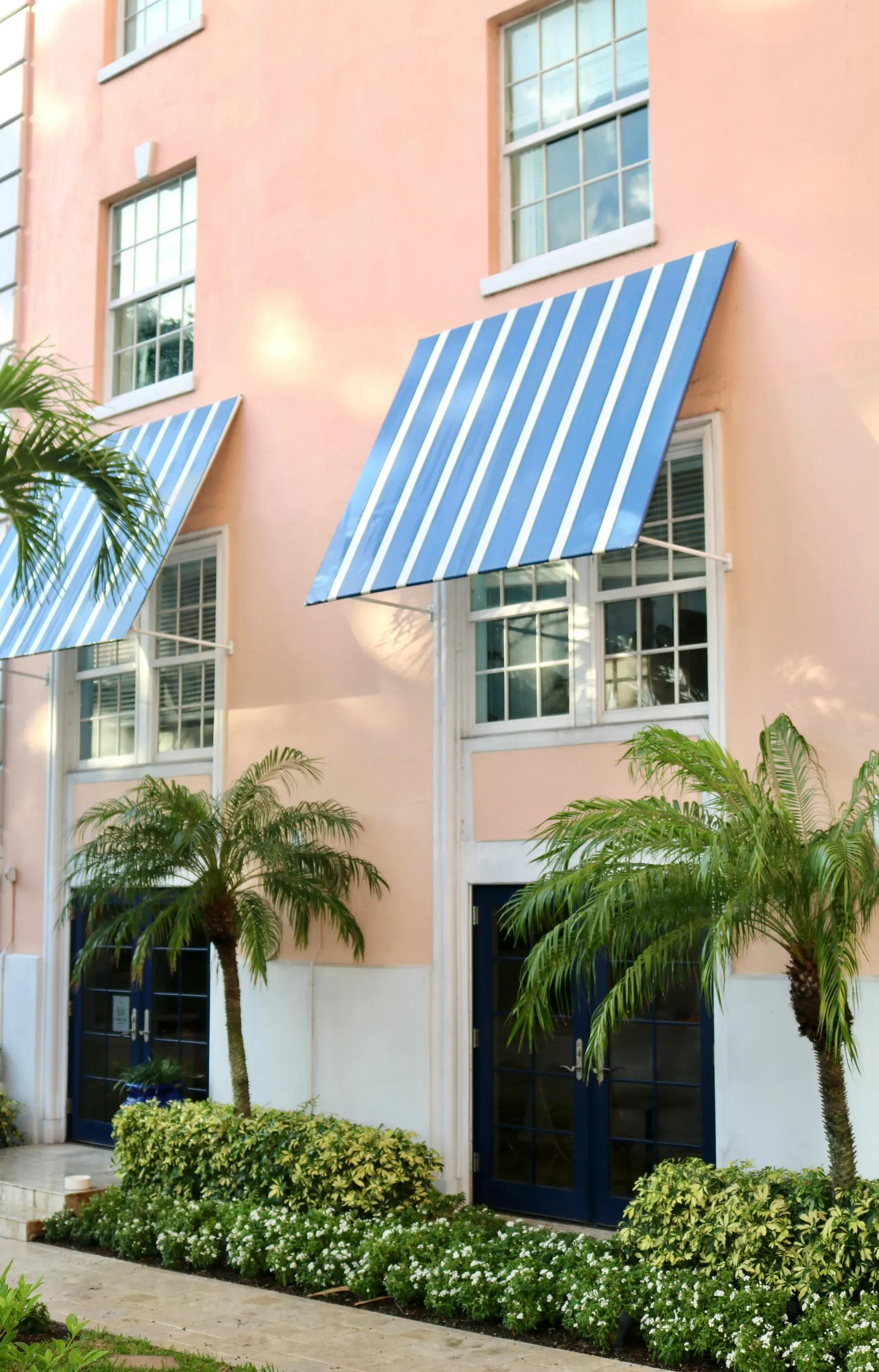 A pink multi-story building facade featuring blue and white striped awnings above windows, surrounded by palm trees and green hedges.