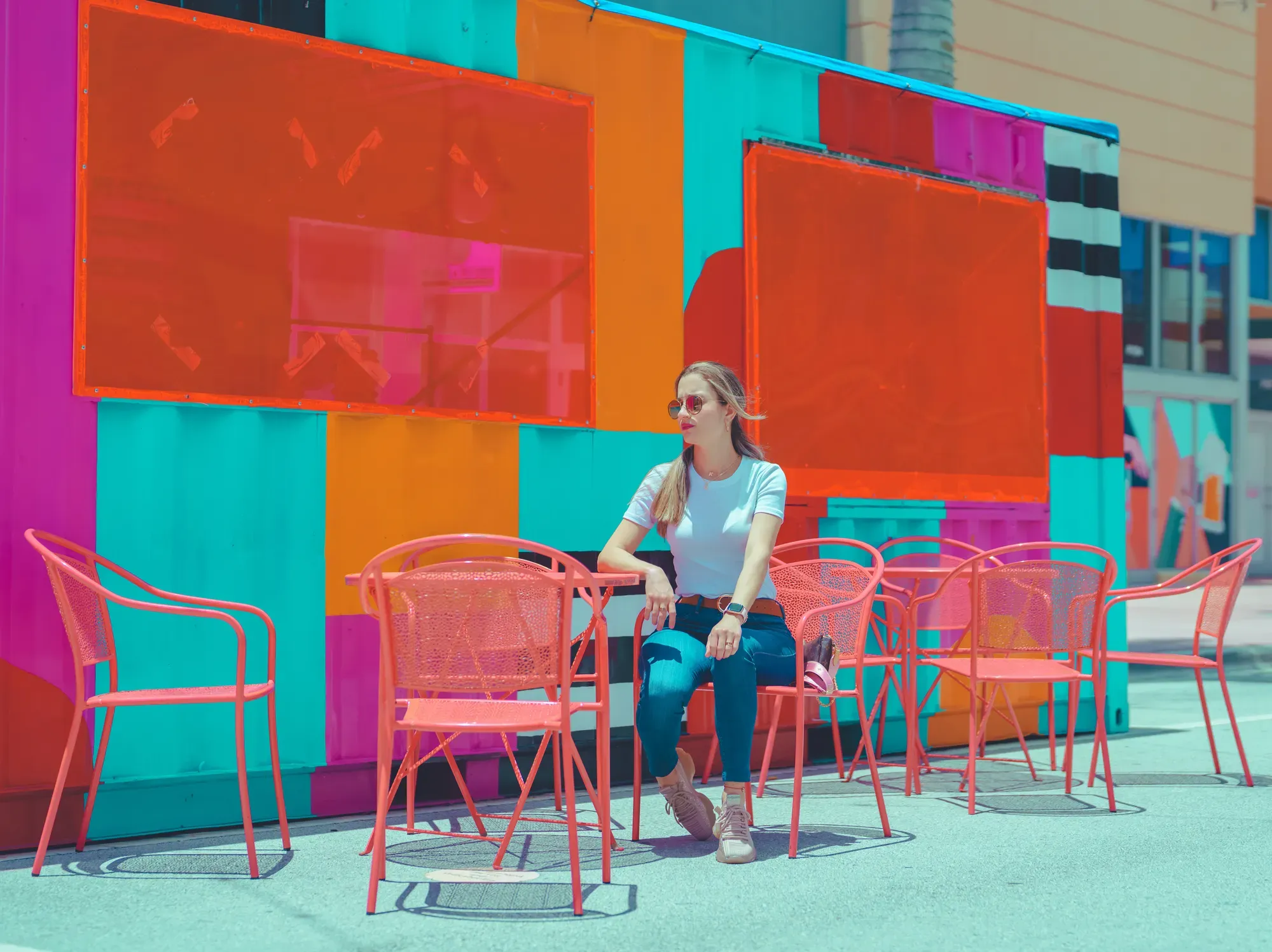 Woman seated at a pink outdoor café table under a bright, colorful geometric wall mural on a sunny day.
