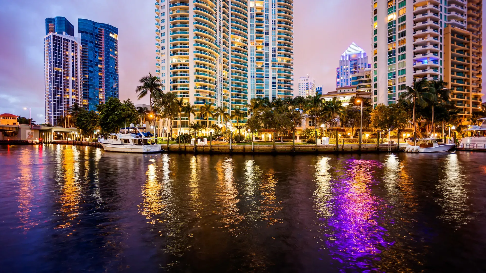 Nighttime city skyline with brightly lit skyscrapers reflecting colorful lights on the calm waterfront, framed by palm trees and docked boats.